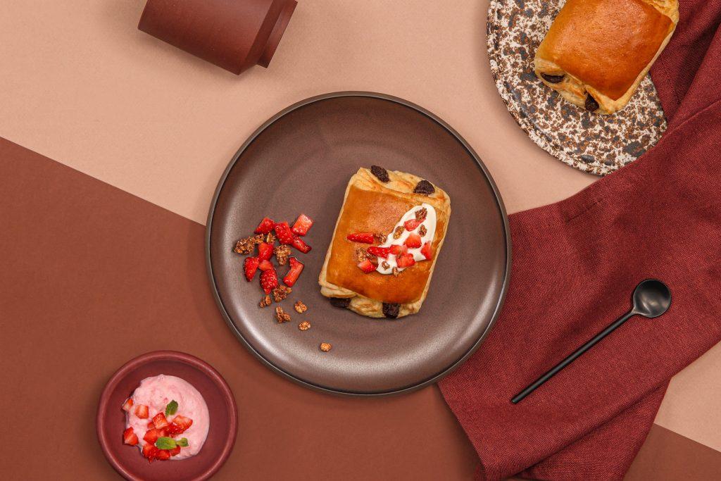Une mise en scène de deux pains au chocolat La Boulangère avec des fraises et du muesli, dans une assiette au centre de l'image