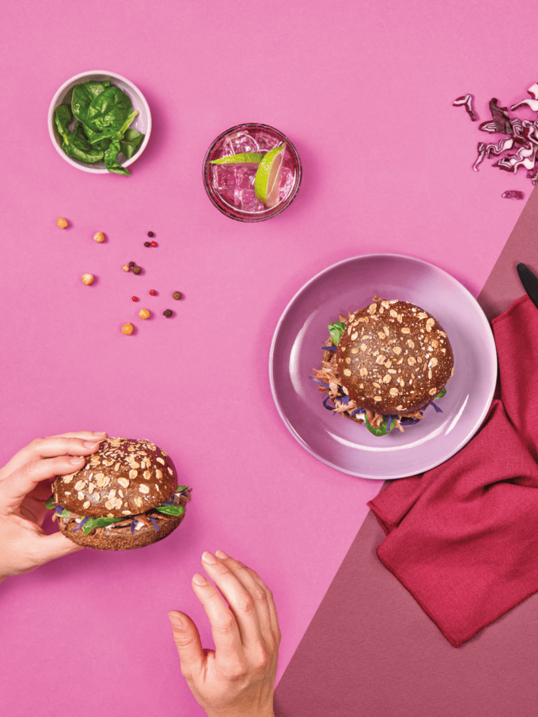 Photo de deux pains burger Seigle et flocons de blé cuisinés. L'un d'eux est tenu par une main. Il y a également des feuilles de basilic ainsi que du chou rouge et des pois chiches.