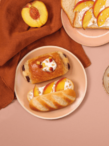 Photo d'un pain au chocolat et d'un pain au lait dans une assiette. Le pain au lait est garnis de fromage blanc et de quartiers de pêche. Une demi-pêche est visible au dessus de l'assiette.